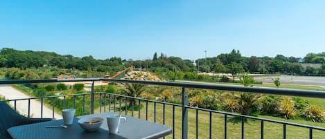 A balcony with a table and chairs, overlooking a landscaped area with trees and a road.