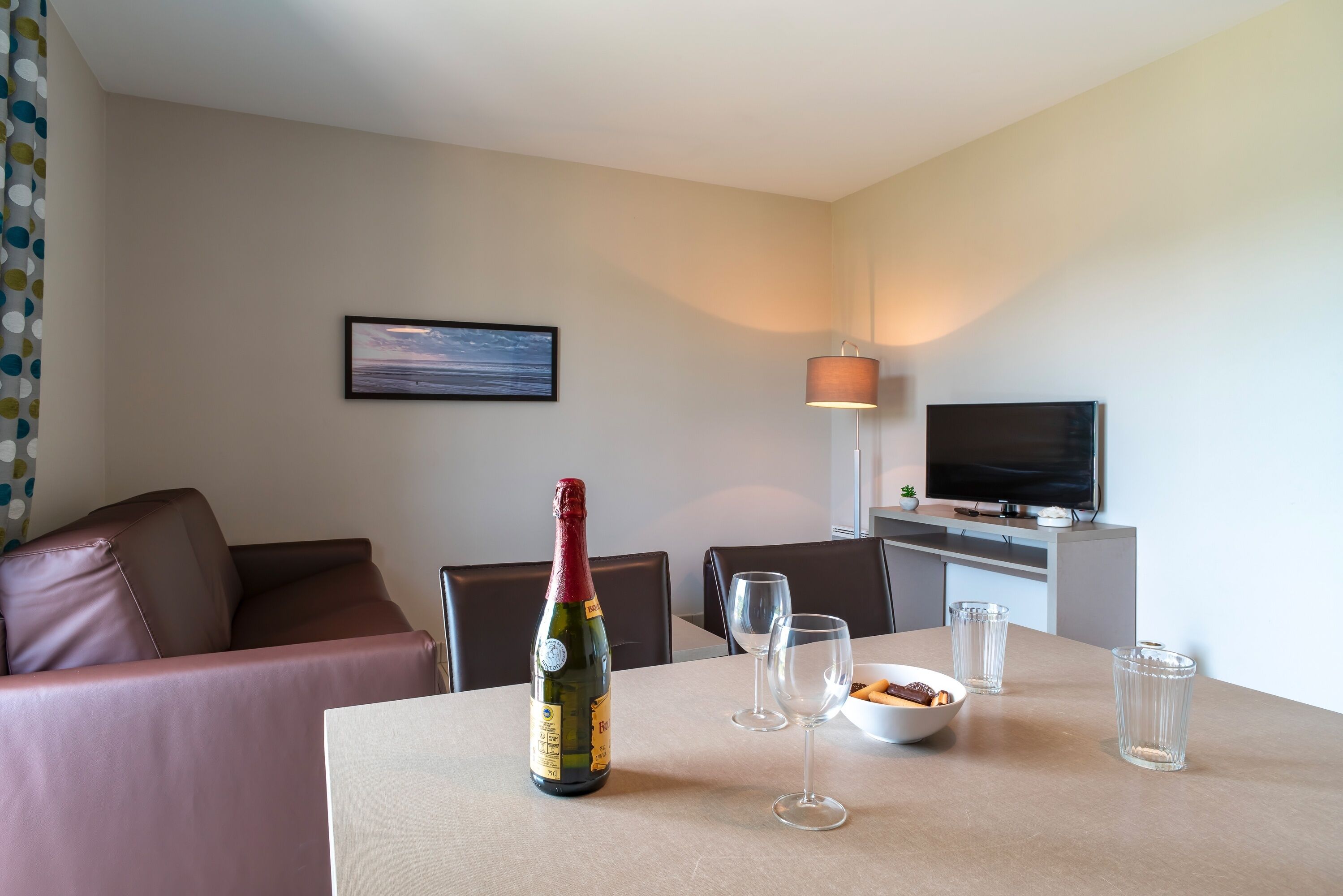 A hotel room with a dining table, a bottle of champagne, two wine glasses, a bowl of snacks, a TV, and a couch.