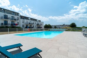 Piscina com espreguiçadeiras, área cercada e um prédio com sacadas ao fundo.