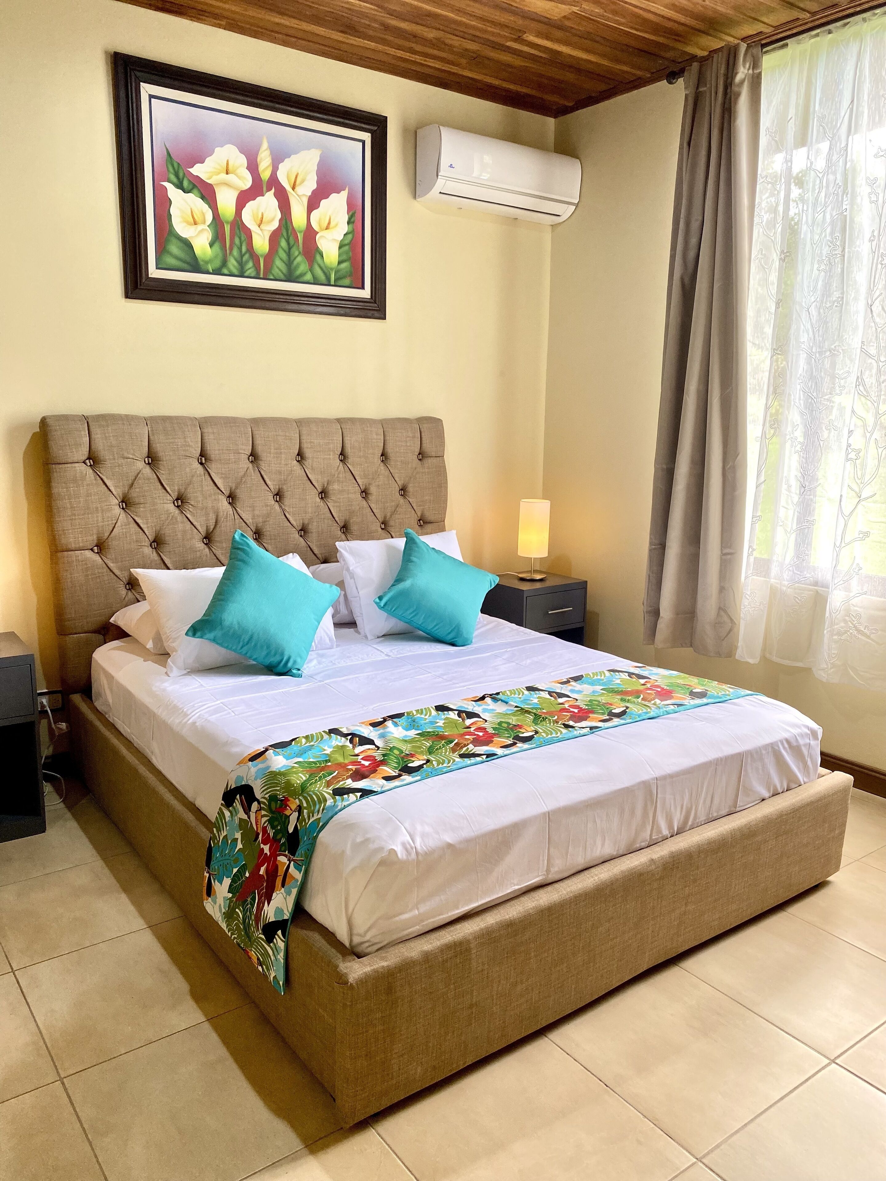 A bedroom with a tufted headboard, a bed with white bedding and teal pillows, a colorful throw blanket, a nightstand with a lamp, an air conditioning unit, and a window with sheer curtains.