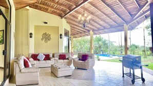 A covered outdoor seating area with a pool in the background. - Mansion Arenal  (La Fortuna)