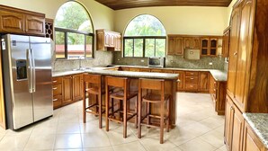 A kitchen with wooden cabinets, a stainless steel refrigerator, a granite countertop, and a large island with bar stools. - Mansion Arenal  (La Fortuna)