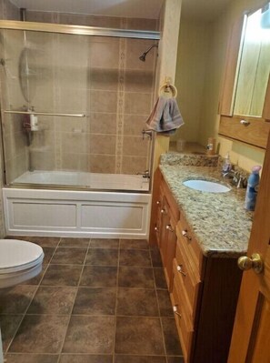 A bathroom with a glass-enclosed shower, a bathtub, a sink with a granite countertop, and wooden cabinets. - Larsen Farm Secluded Farmhouse with Hotub
 (Wauzeka)