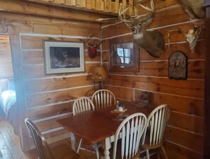 A wooden cabin interior with a dining table and chairs, framed pictures, and mounted animal heads on the walls.