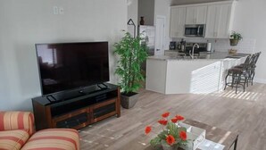 Uma sala de estar moderna com televisão de tela plana, um sofá, uma mesa de jantar com um vaso de flores e uma cozinha ao fundo.