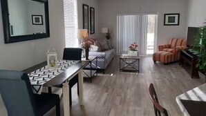 Uma sala de estar com uma mesa de jantar, sofá e poltronas, um ventilador de teto e uma janela grande com persianas.