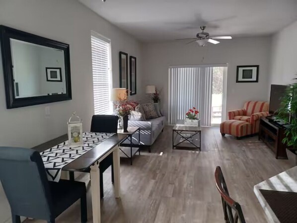 Un salon comprenant une table à manger, un canapé et des fauteuils, un ventilateur de plafond et une grande fenêtre avec des stores.
