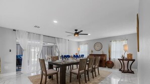 A dining area with a marble floor, a large wooden table, and chairs. There is a ceiling fan, a clock on the wall, and a lamp on a side table.