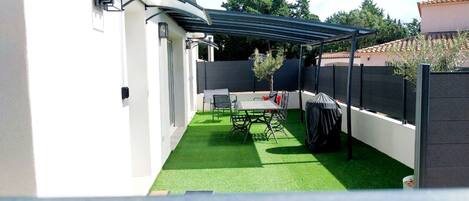 Een overdekt terras met kunstgras, een tafel, stoelen en een barbecue.