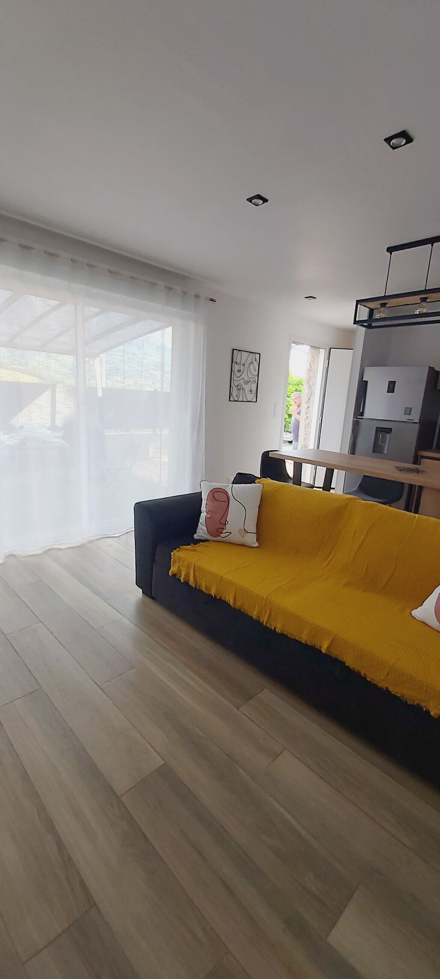 A modern living room with a yellow sofa, wooden flooring, and a large window with sheer curtains.
