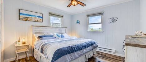 A bedroom with a wooden floor, a bed with a headboard, a ceiling fan, and a window with a view of the outdoors.