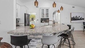 A modern kitchen with granite countertops, white cabinetry, and a central island with bar stools. - The Coral Reef Oasis (Cape Coral)