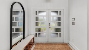 A hallway with a white door featuring a grid pattern, a bench, a woven light fixture, and a textured rug. - The Coral Reef Oasis (Cape Coral)