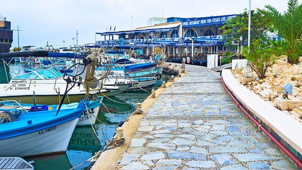 Un port de plaisance avec des bateaux amarrés, une promenade pavée et un bâtiment à la façade bleue qui fait de la publicité pour les fruits de mer.