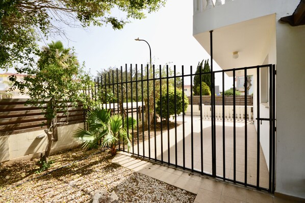 A gated courtyard with a paved pathway, surrounded by a fence and various plants.