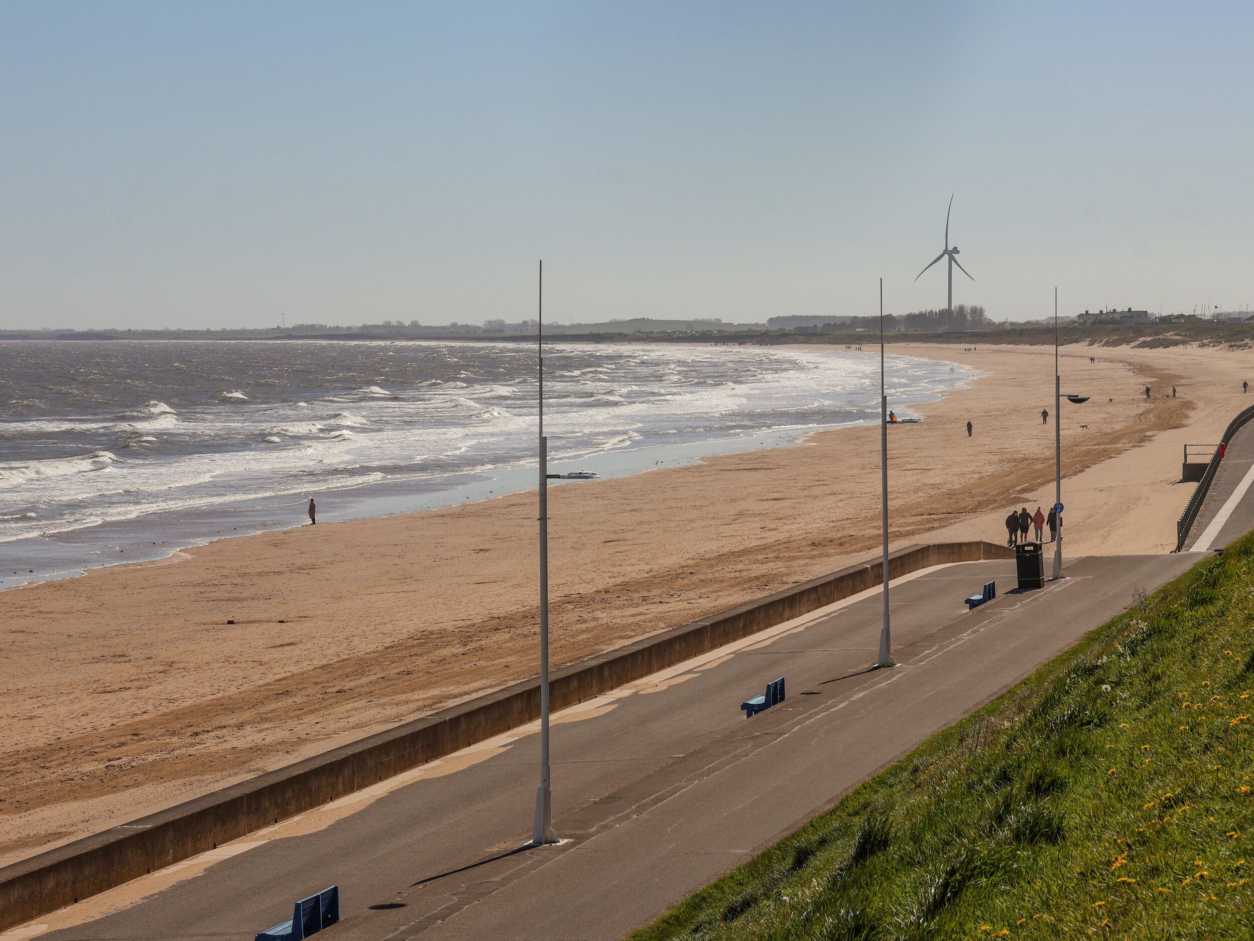 A coastal road with a beach on one side, a wind turbine in the distance, and a clear sky.