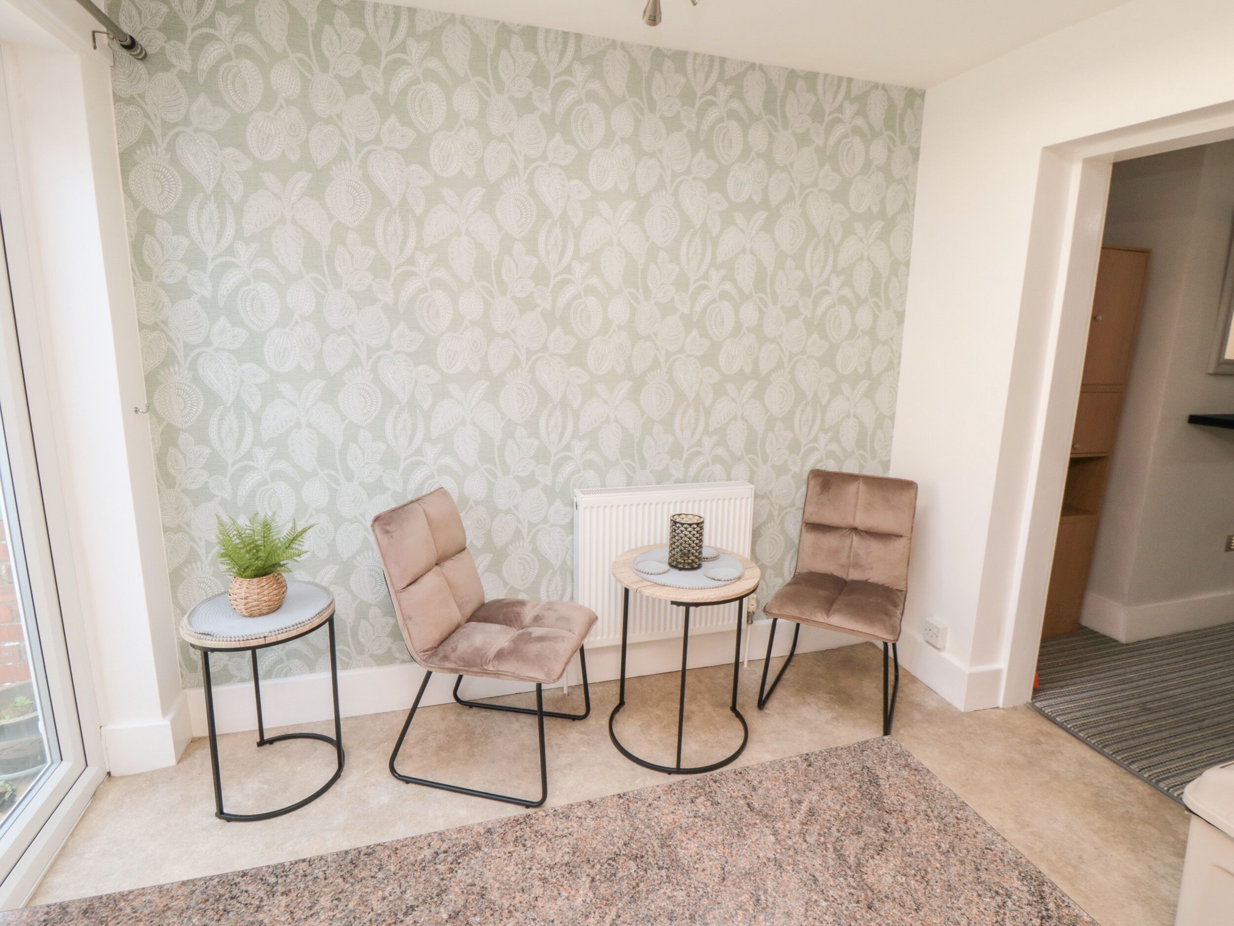 Una habitación con papel tapiz estampado, una pequeña mesa redonda con una planta, dos sillones con cojines y un radiador.