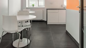 A modern kitchen with white cabinets, a white dining table, and chairs. There is a window with blinds and a small plant on the windowsill. - ESPERANTO HOME (Katowice)