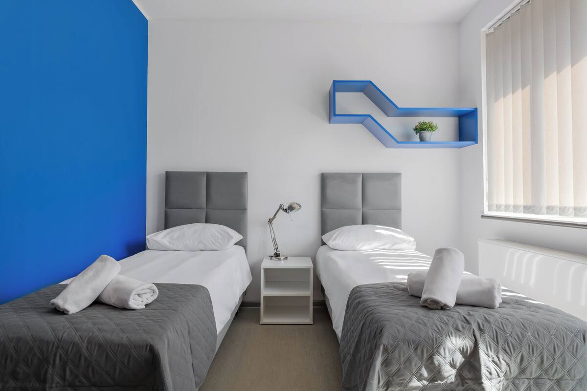 A modern hotel room with two beds, a nightstand, a desk lamp, and a blue shelf with a plant.