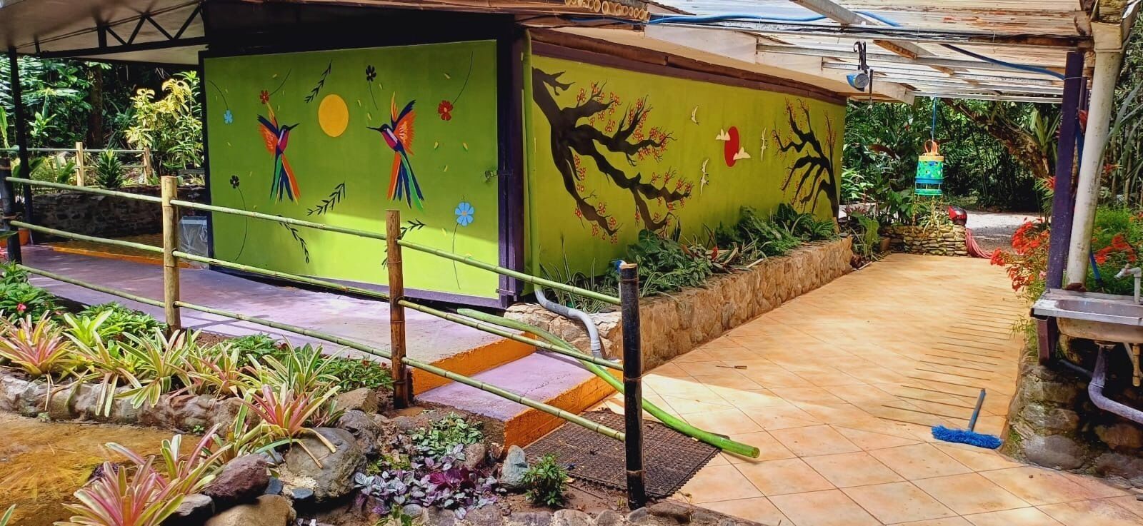 A covered outdoor area with a wooden pathway, a small stream, and colorful murals on the walls.