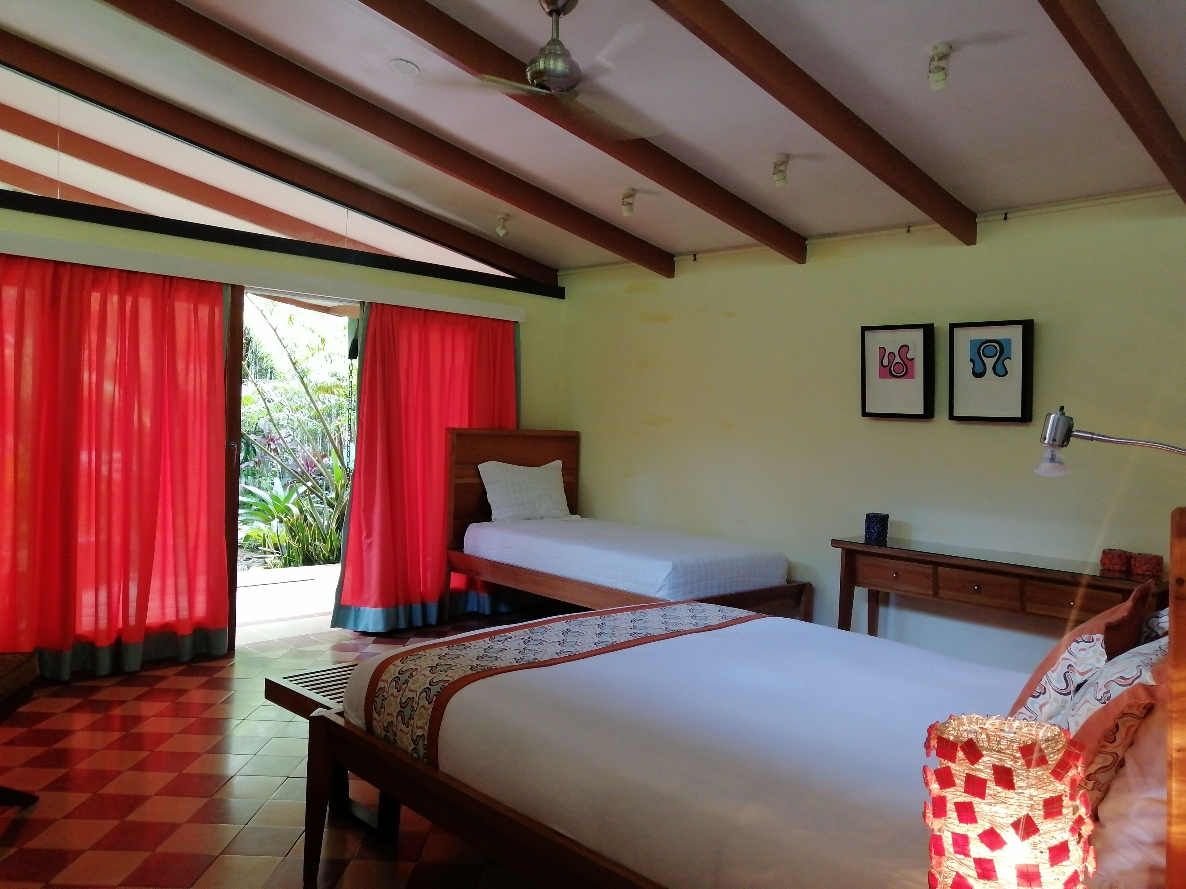 Quarto com cama, escrivaninha e cadeira. Possui duas janelas com cortinas vermelhas.