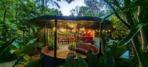 An outdoor dining area with a covered seating arrangement and hanging lights, surrounded by lush greenery. - Chirripo Art Hotel (Rivas)