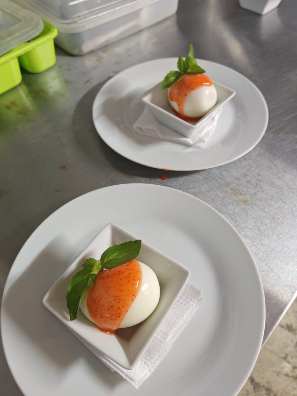 Two white plates with a dessert consisting of a white sphere and orange sauce, garnished with green leaves, on a stainless steel surface.