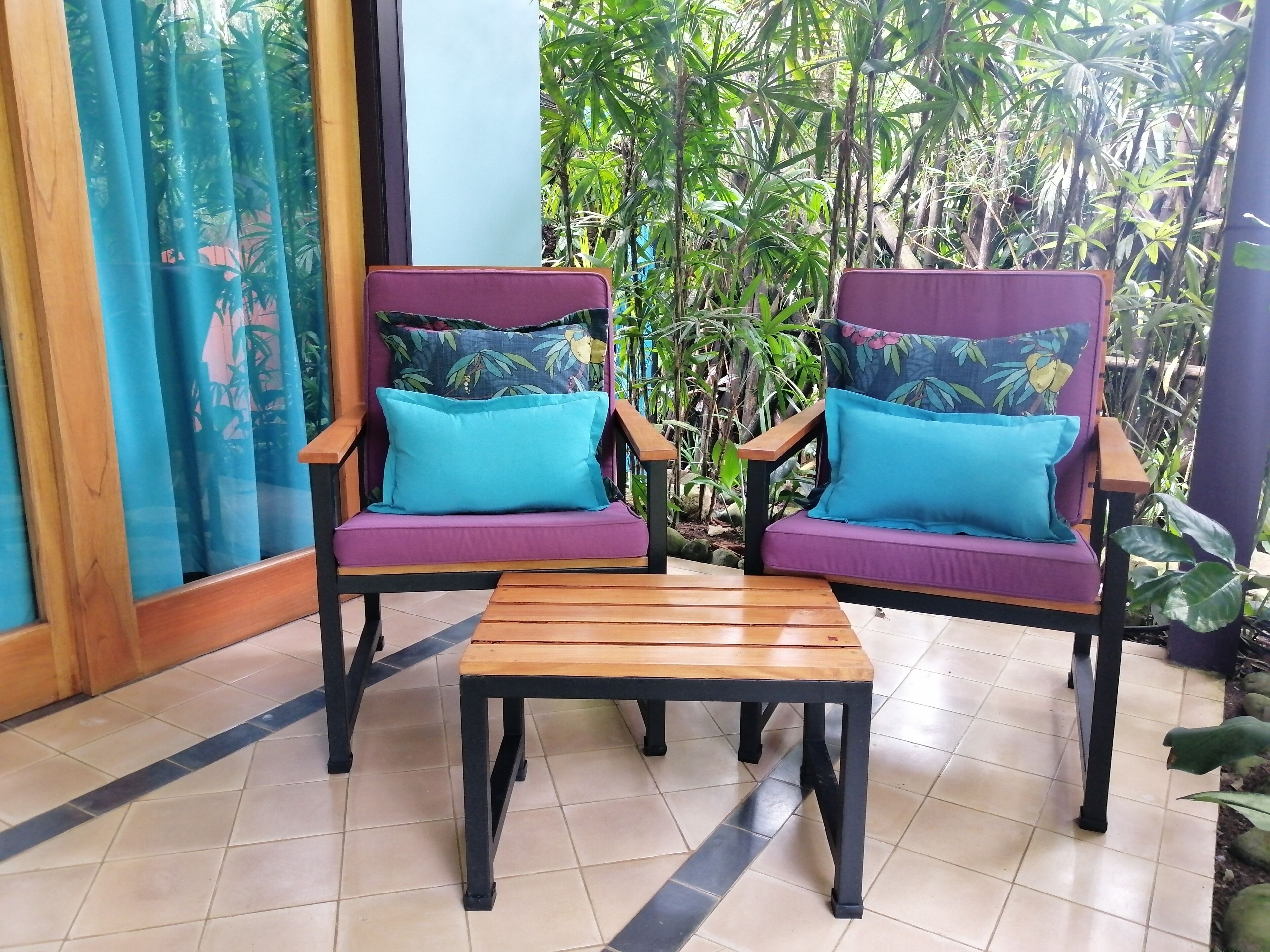 Dos sillones morados con cojines color turquesa y una mesa de madera en una habitación con suelo de baldosas y vistas a un follaje denso.