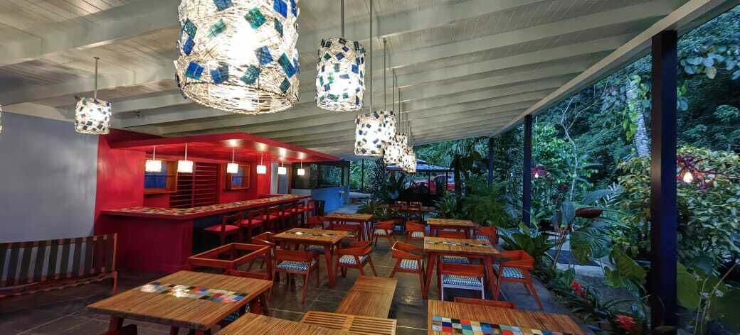Un restaurant intérieur avec des tables et des chaises en bois, des luminaires décoratifs suspendus et une vue sur la verdure à l’extérieur.
