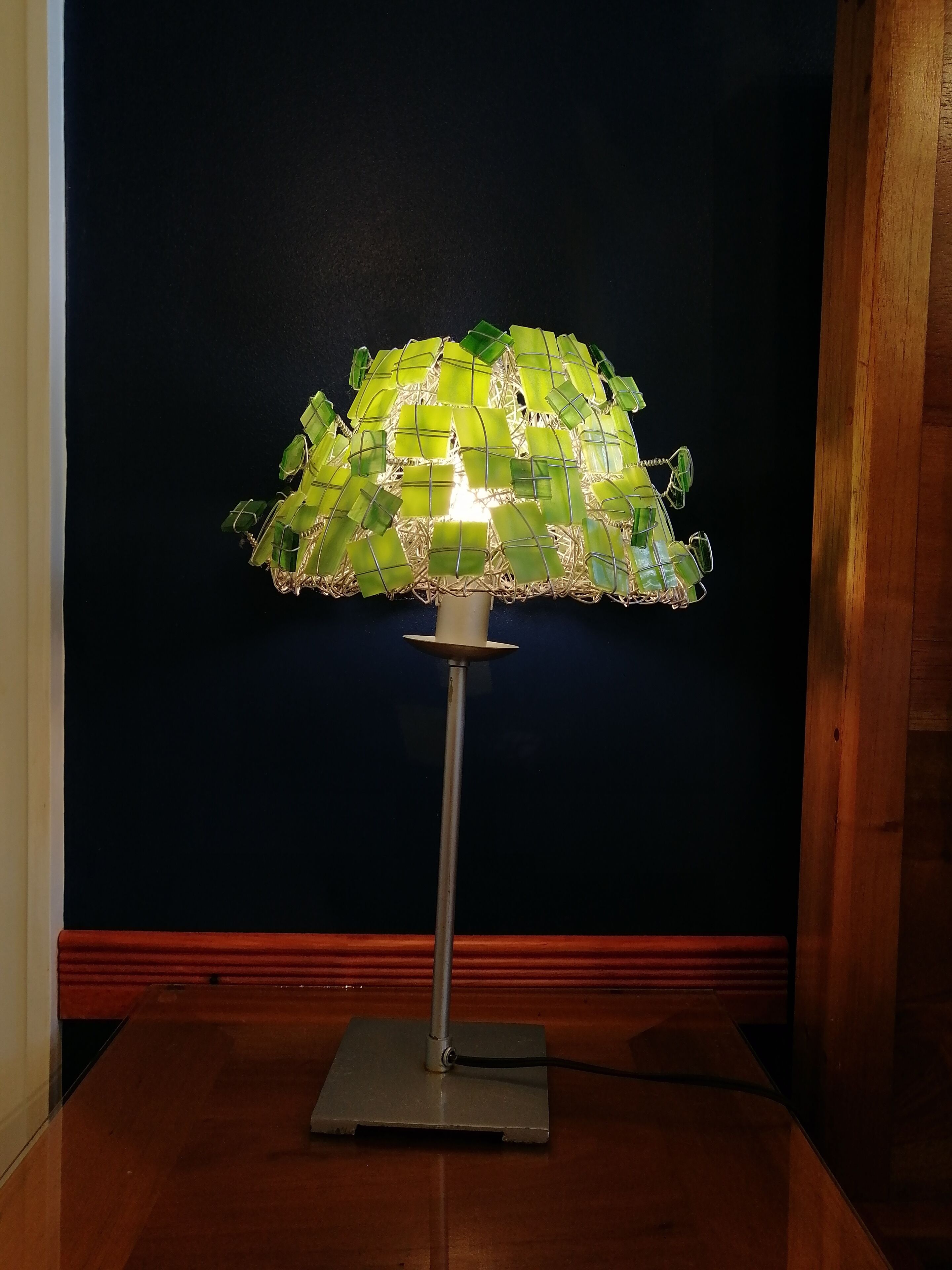 Una lámpara de pie con pantalla de vidrio verde sobre una mesa de madera, frente a una pared oscura.