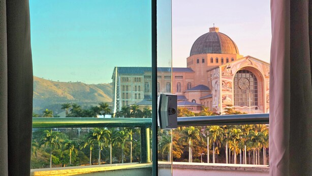 A view from a window with a metal railing, overlooking a building with a dome and arched windows, palm trees, and a hilly landscape. - MAPP HOTEL (Aparecida)