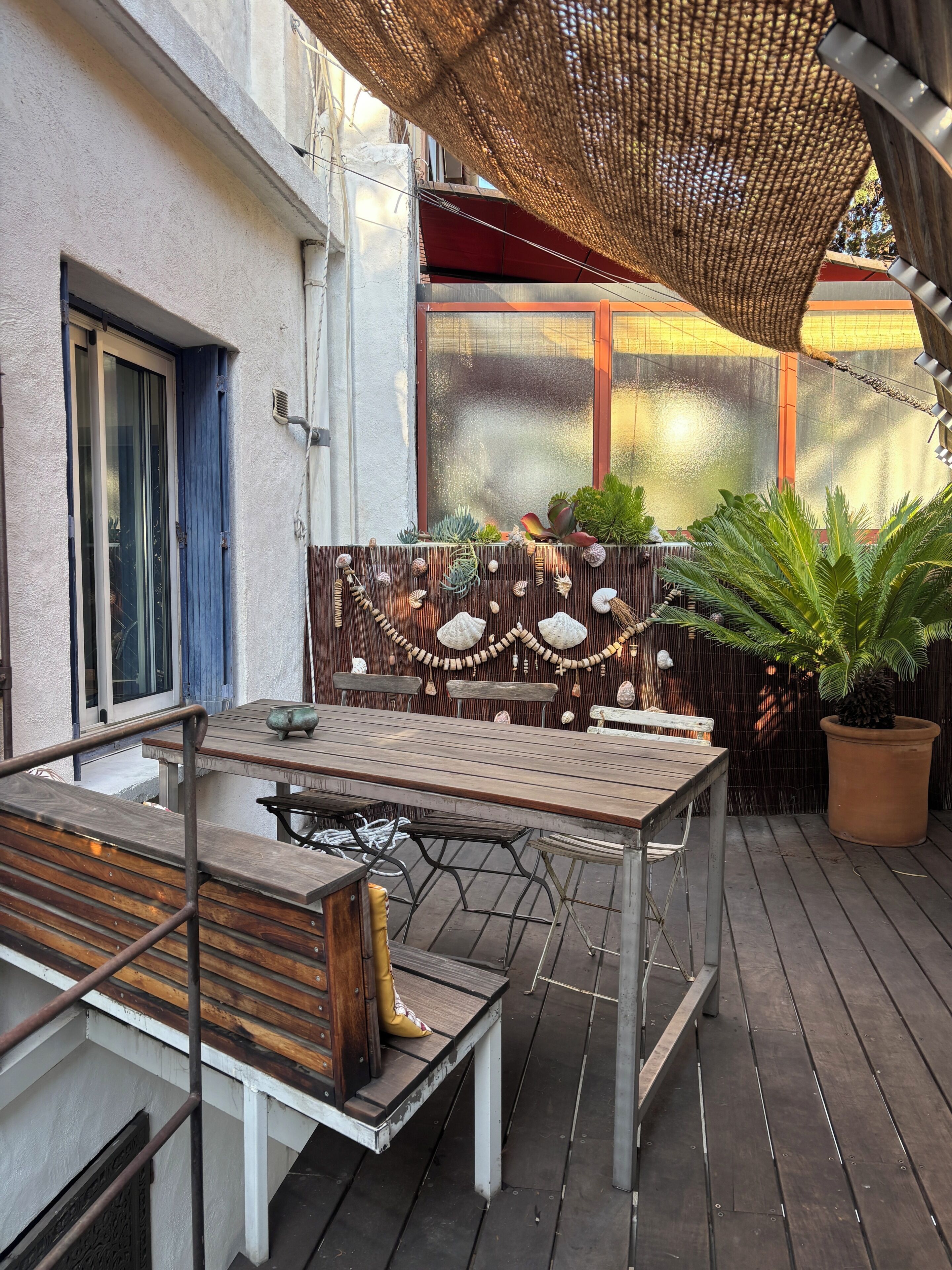 An outdoor patio with a wooden table and benches, surrounded by potted plants and a decorative wall.