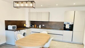 A modern kitchen with a wooden dining table, white chairs, a washing machine, a microwave, and a refrigerator.