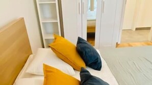 A bed with white bedding and two pillows, one yellow and one dark blue, in a room with a wooden headboard and a white wardrobe.