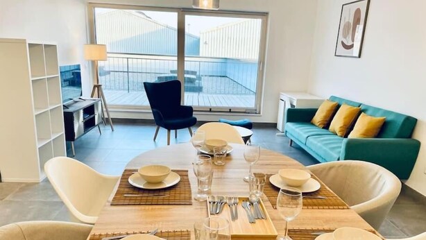 A dining area with a table set for four, a blue chair, a teal sofa with yellow cushions, and a large window with a view of a building.