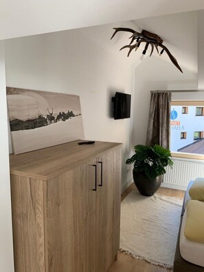 A room with a wooden cabinet, a mounted TV, a painting of deer, a potted plant, and a view of a building outside.