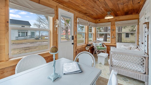 Una habitación acogedora con paredes y techo de madera, una mesa blanca con lámpara, muebles de mimbre y ventanas amplias que dan a vistas del exterior.