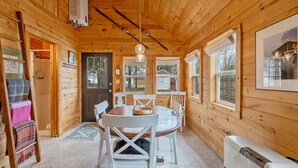 Interior de una cabaña de madera con una mesa de comedor, sillas, chimenea y un ventanal grande.