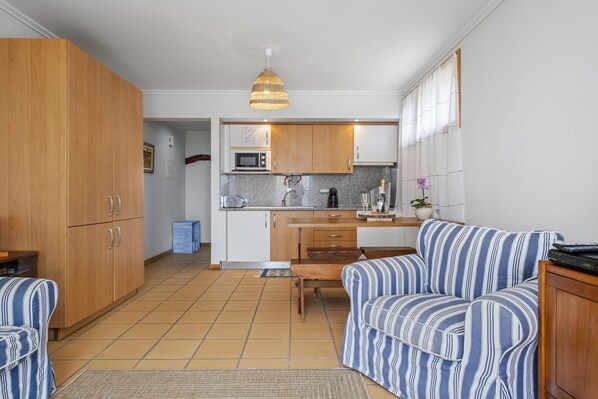Una cocina compacta con armarios de madera, un microondas y un lavamanos. Hay un sofá a rayas y una mesita en la sala.