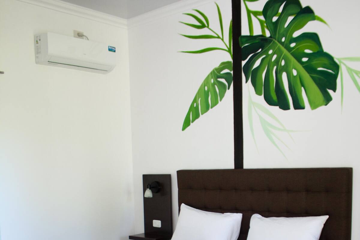 Un lit bien fait, avec une tête de lit marron, un climatiseur mural blanc et une grande plante verte servant de décoration.