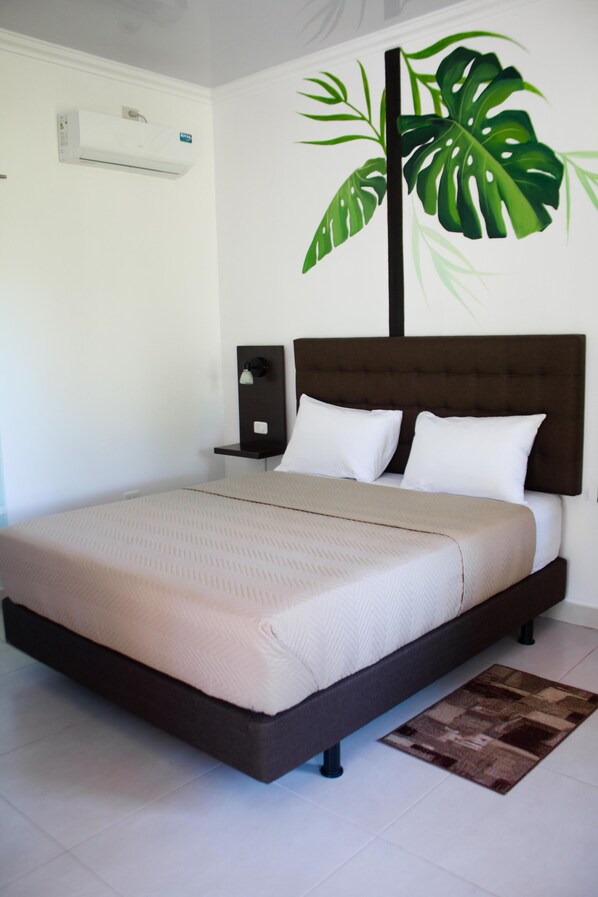 Una cama bien hecha con un cabecero marrón, un aire acondicionado blanco en la pared y una planta verde decorativa grande.