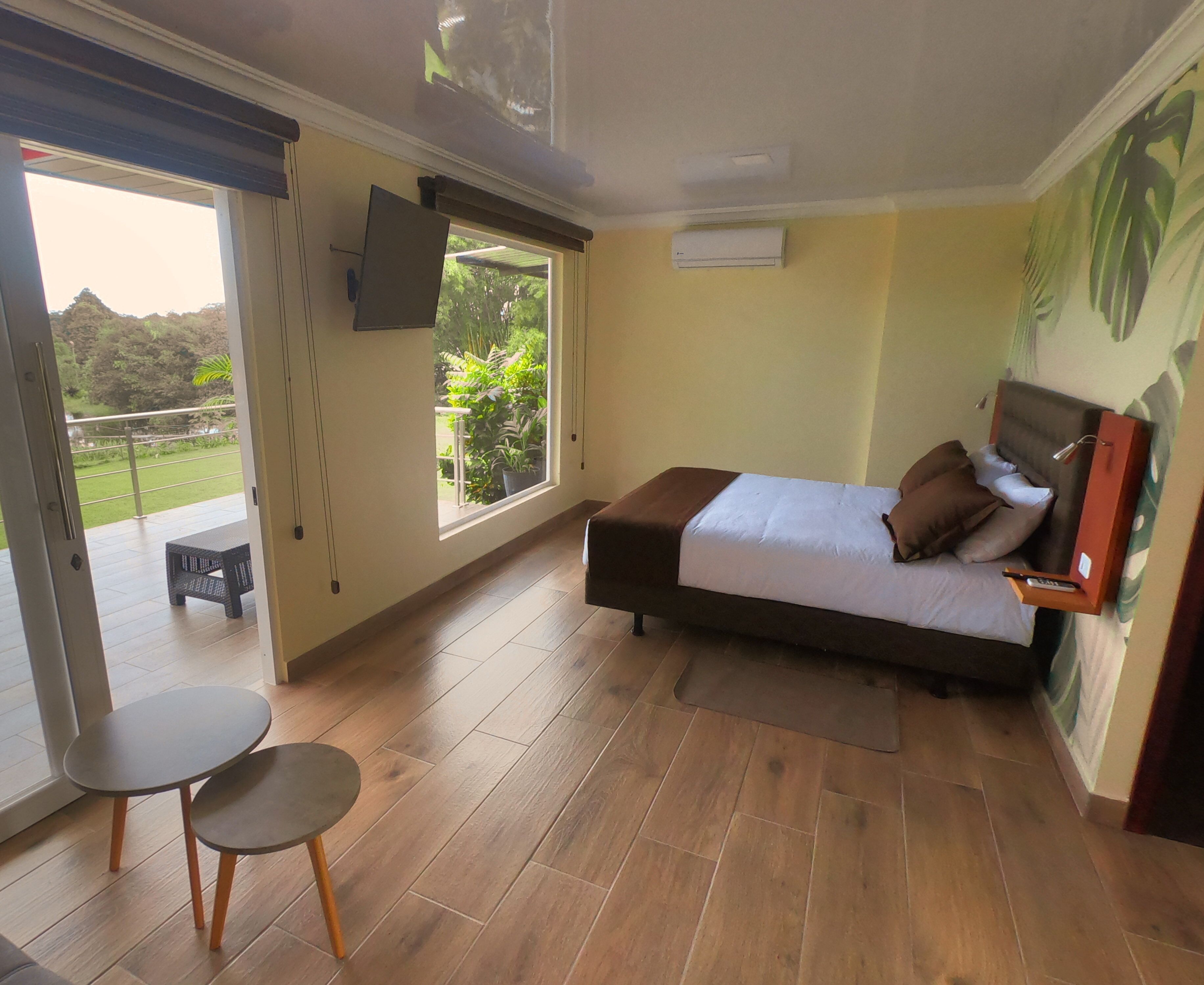 A bedroom with a bed, a TV mounted on the wall, a small table, and a view of the outdoors through a sliding door.