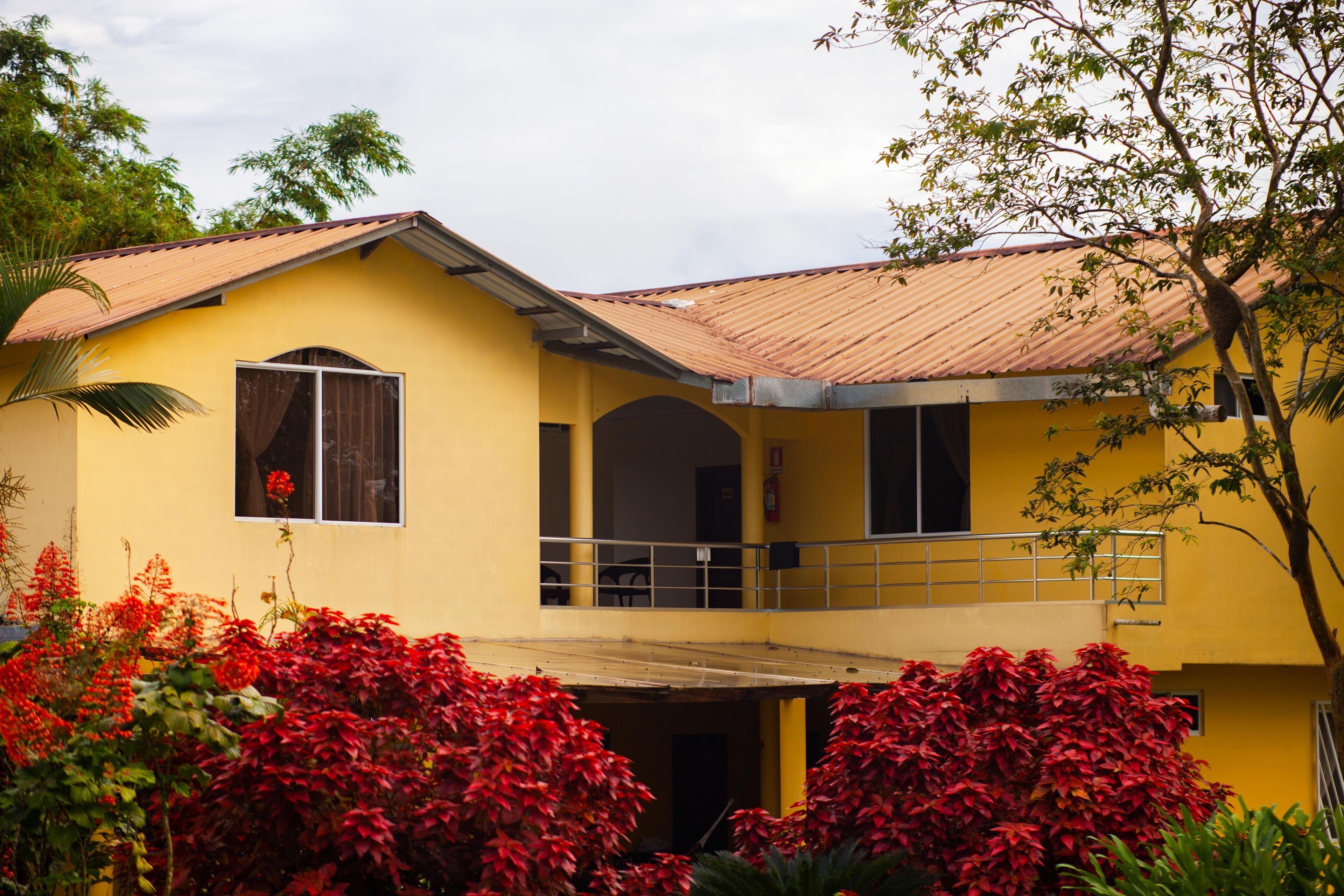 A yellow building with a red roof, a balcony, and red foliage in the foreground.
