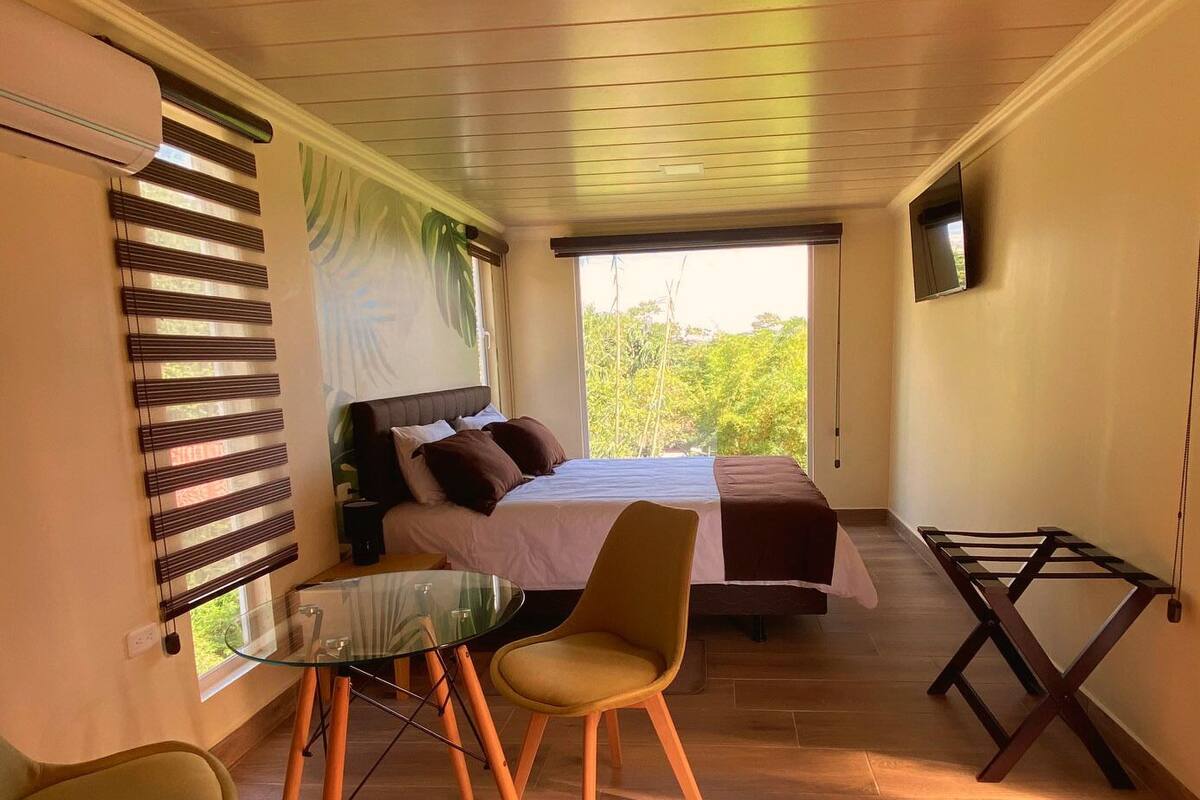 Une chambre à coucher avec un lit, une table en verre, deux chaises, un téléviseur et une fenêtre avec des stores.