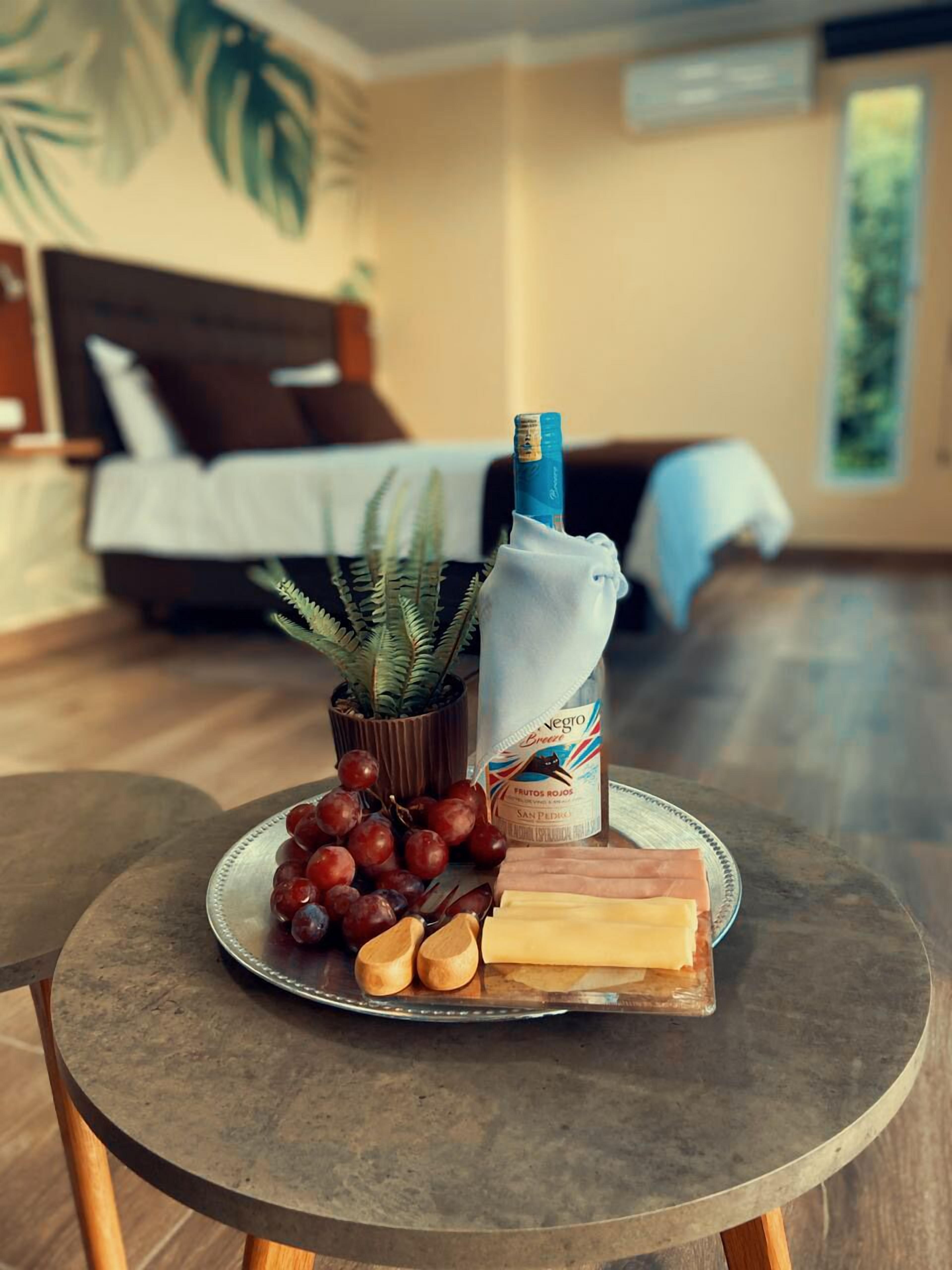 A dining table with a plate of grapes, cheese, and a bottle of juice in a room with a bed and a plant in the background.
