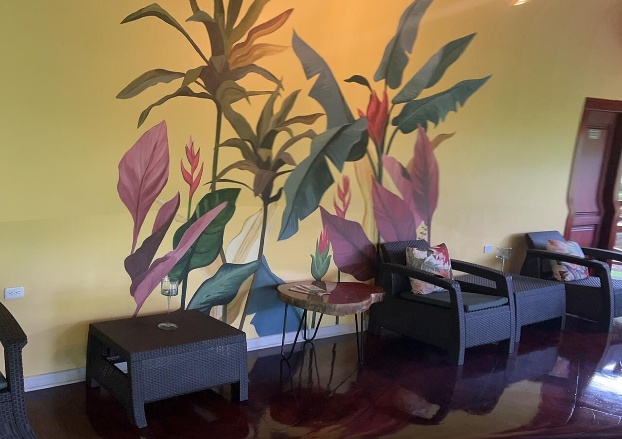 A room with a large wall mural of tropical plants, a round table with a glass, and two wicker sofas.
