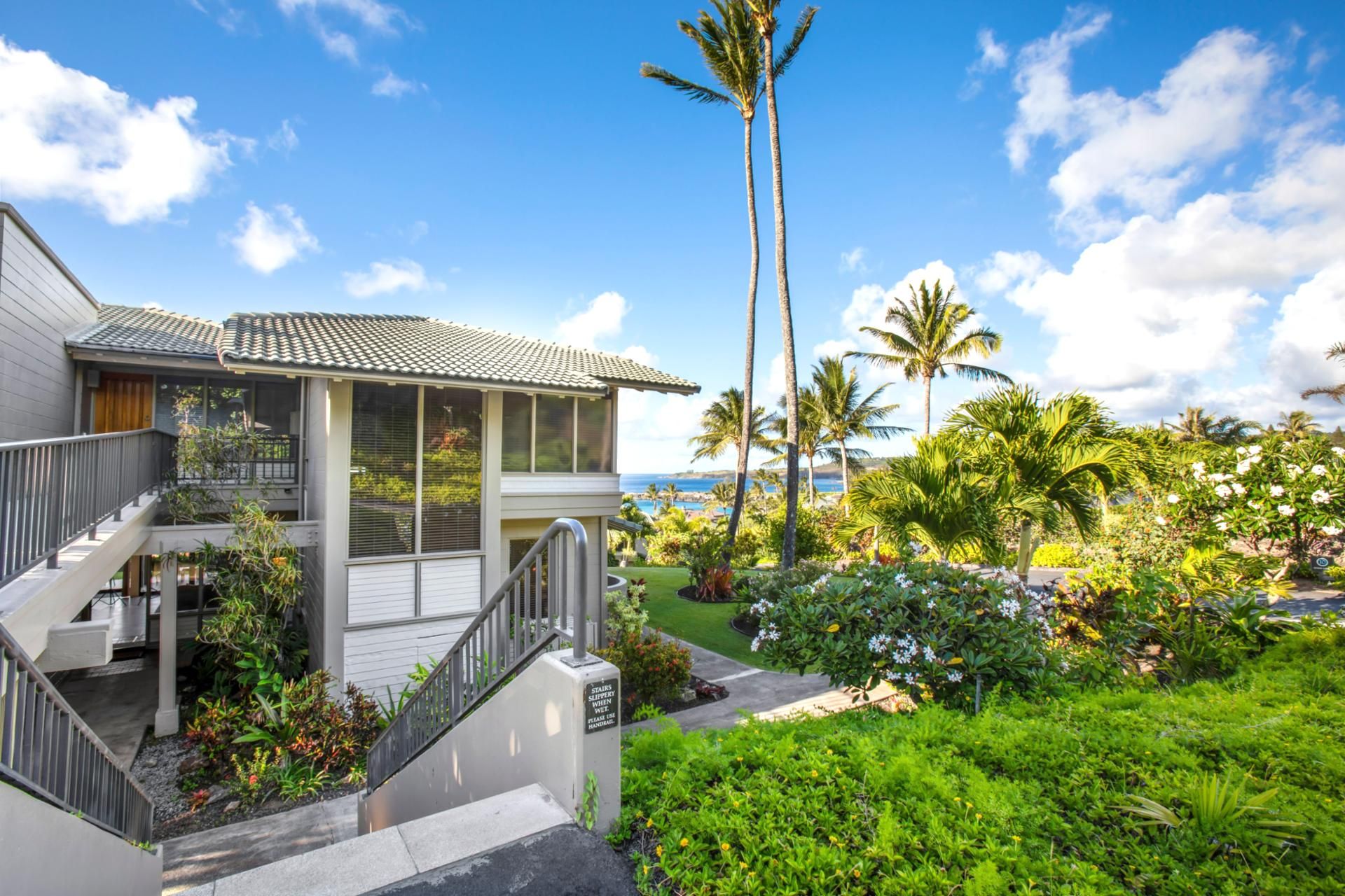 A two-story house with a balcony, surrounded by lush greenery and palm trees, overlooking a scenic view.