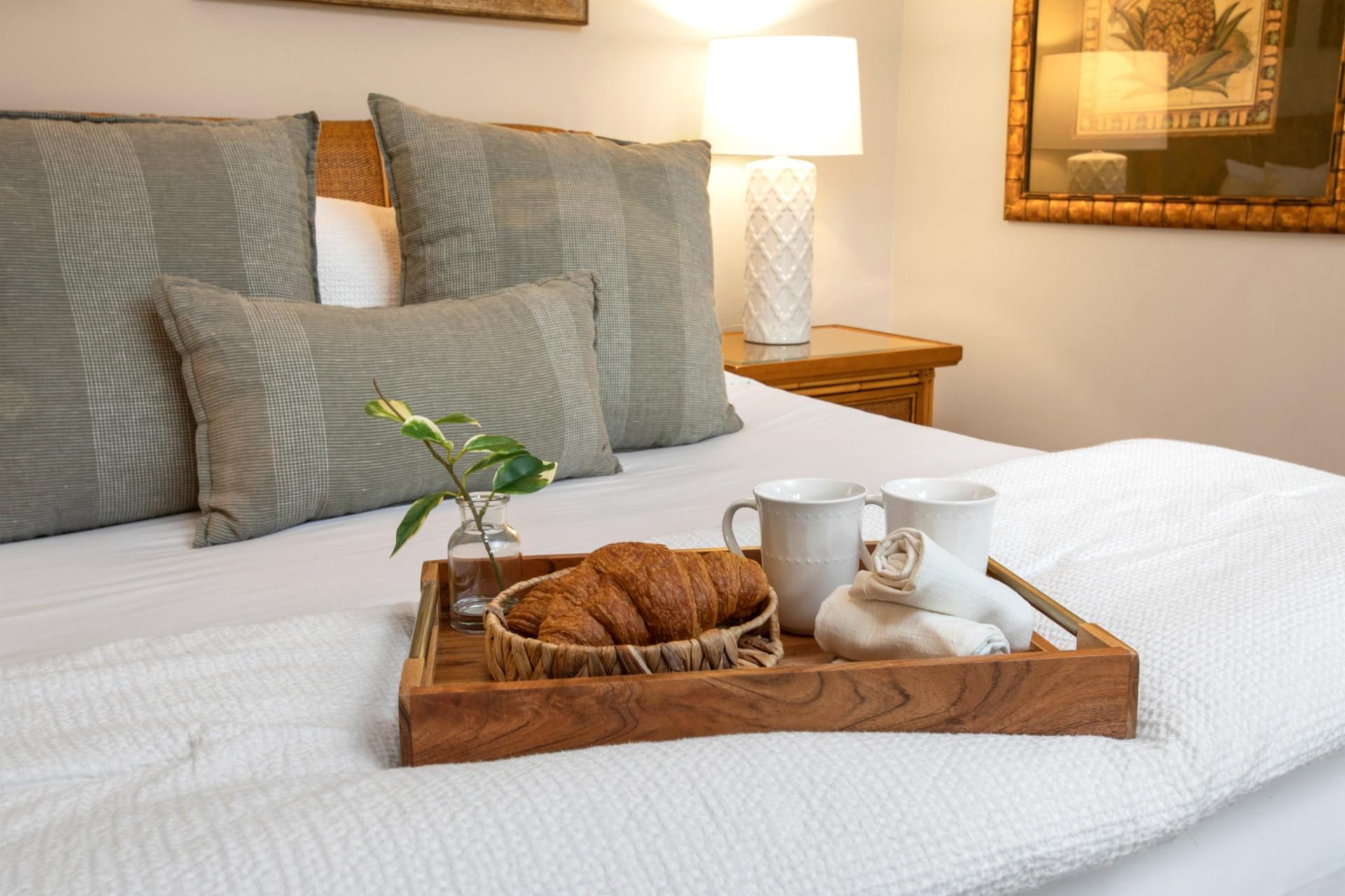 A neatly made bed with a wooden tray containing croissants, a vase with a plant, and two white mugs.