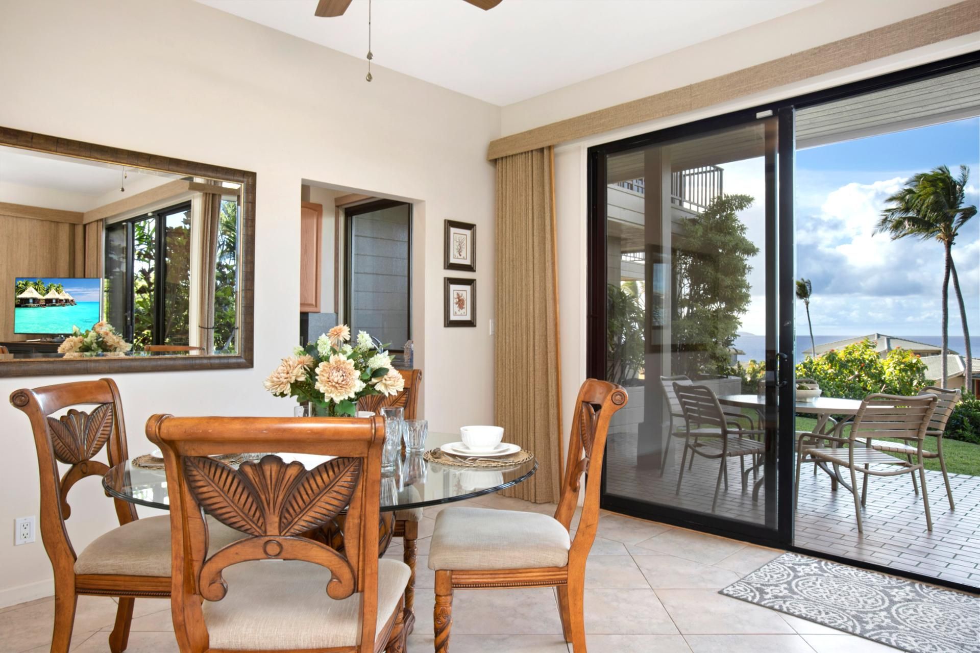 A dining area with a glass table, two wooden chairs, a vase of flowers, and a television displaying a tropical scene.