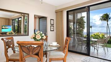 A dining area with a glass table, two wooden chairs, a vase of flowers, and a television displaying a tropical scene.